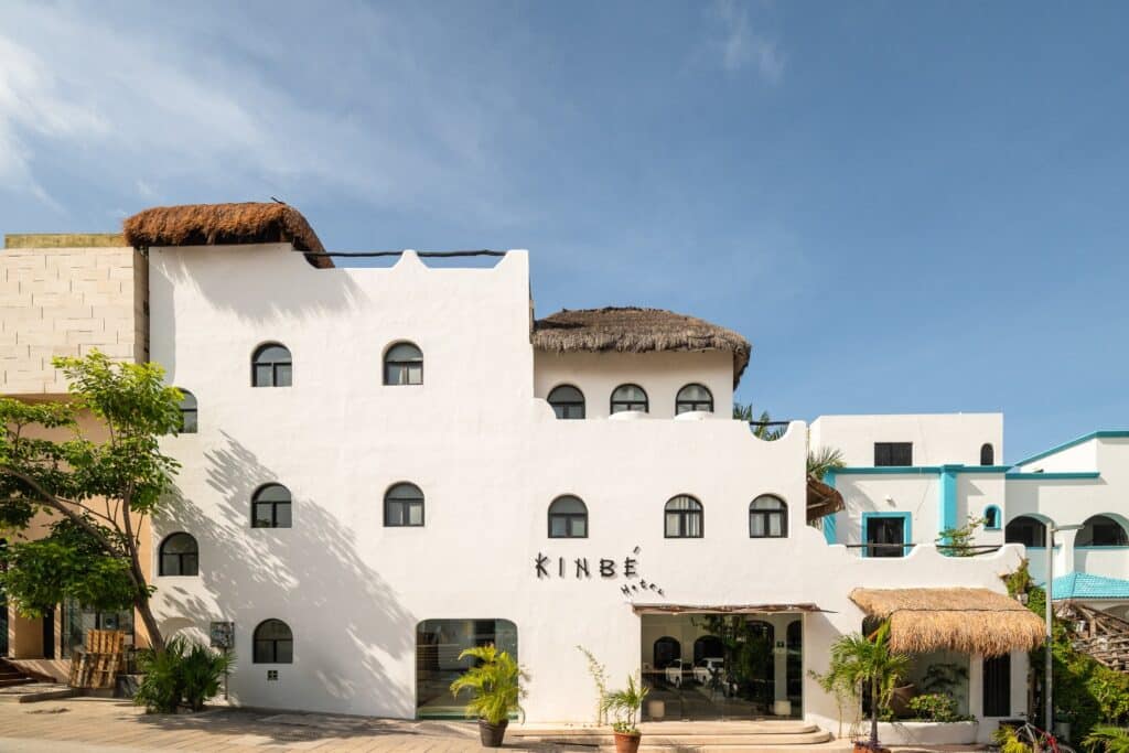 A white building with arched windows and thatched roofs, part of the Kinbe Hotel in Playa del Carmen's Centro area, featuring a modern colonial design against a blue sky.