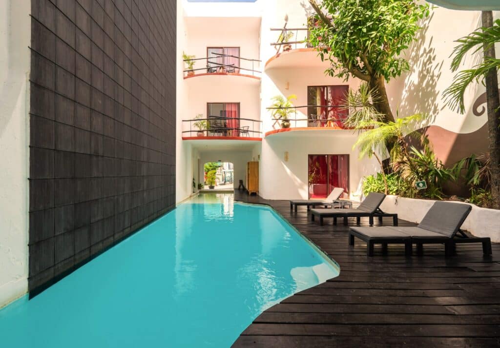 A cozy courtyard at Kinbe Hotel in Centro Playa Del Carmen features a small swimming pool, surrounded by wooden decking, lounge chairs, and lush tropical plants.