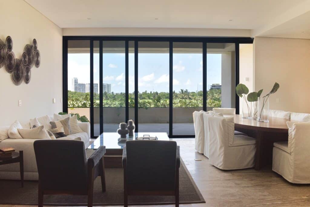 Living room in La Vela 412, Puerto Cancun, featuring a spacious area with white couches, dark chairs, a glass coffee table, and large sliding doors leading to a balcony with a green view.