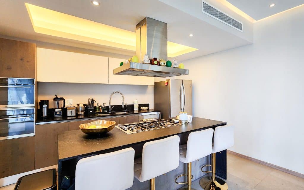A modern kitchen in Paradox 24 A, Santa Fe, Mexico City, featuring a sleek black countertop, built-in appliances, and four white upholstered bar stools at the island.