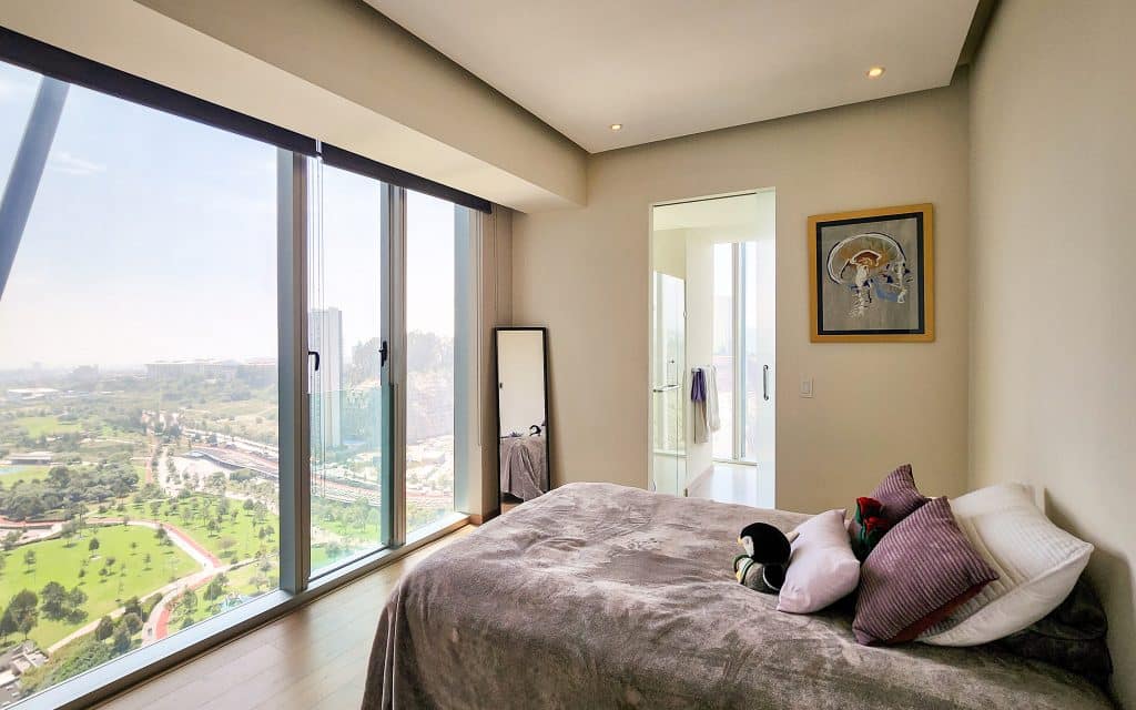 A bedroom in Paradox 24 A, Santa Fe, Mexico City, with a large window offering views of a green park and cityscape, decorated with stuffed toys and colorful pillows.