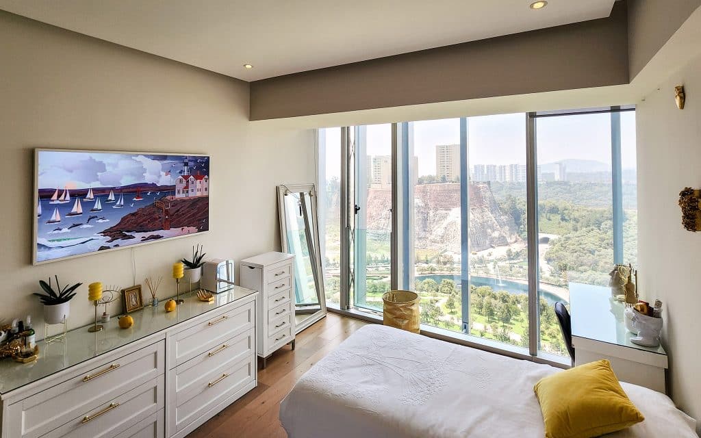 A bedroom in Paradox 24 A in Santa Fe, Mexico City, featuring a large window with a view of the cityscape, a white dresser decorated with plants and ornaments, and a bed with a yellow pillow.