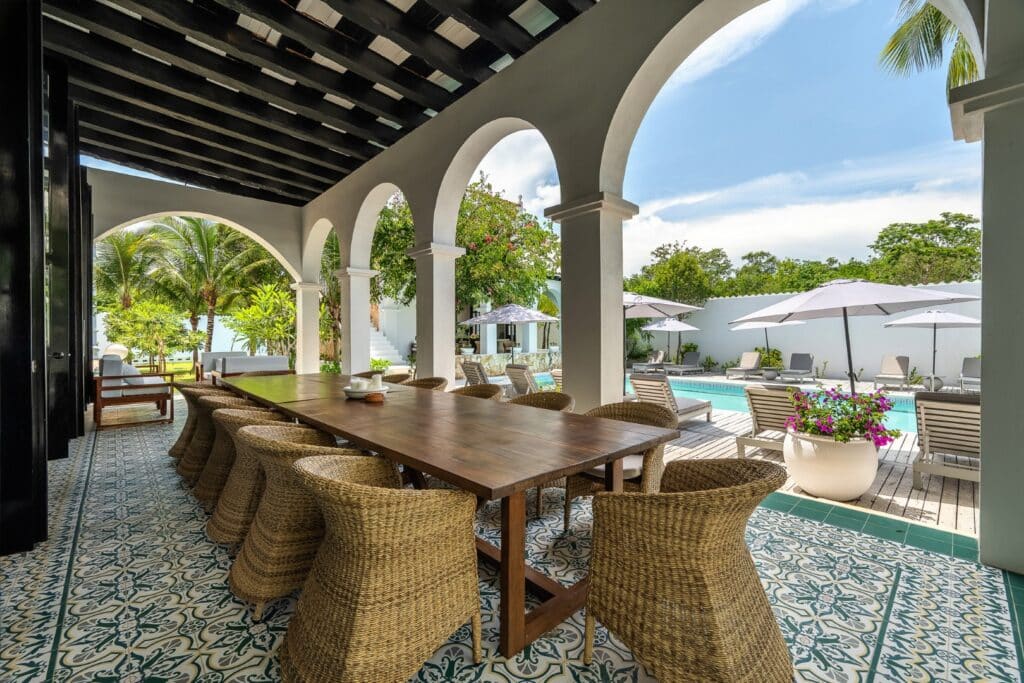 Covered veranda with arched white columns, a large wooden dining table, pool area with loungers and umbrellas, lush greenery