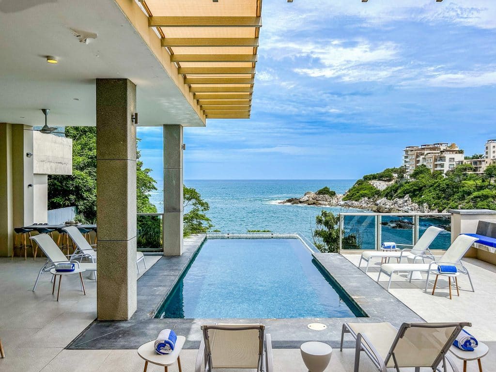 An outdoor view from Villa Montecarlo in Playa Arrocito, Huatulco, featuring a swimming pool with a scenic ocean and rocky coastline in the background, surrounded by modern buildings and lush greenery.