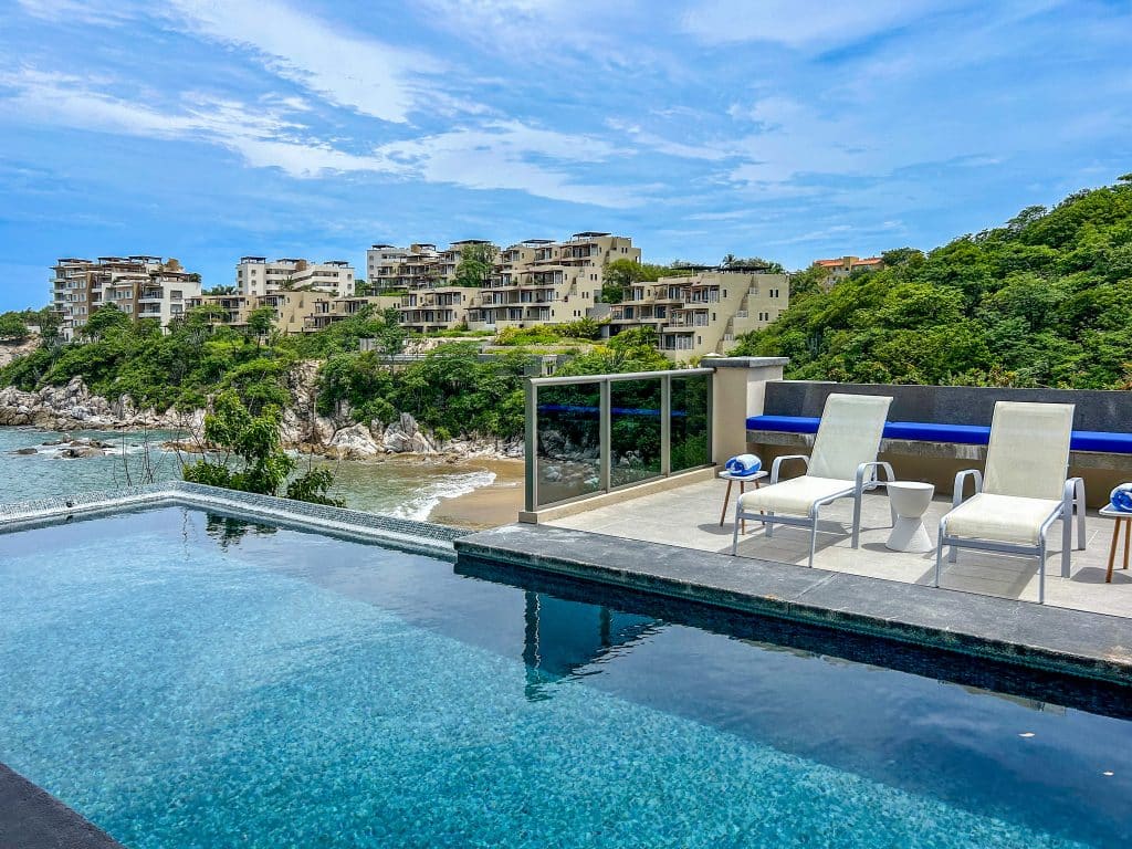 A beachfront swimming pool with lounge chairs and a scenic view of Villa Montecarlo in Huatulco's Playa Arrocito area, overlooking neighboring modern apartment buildings and lush greenery.