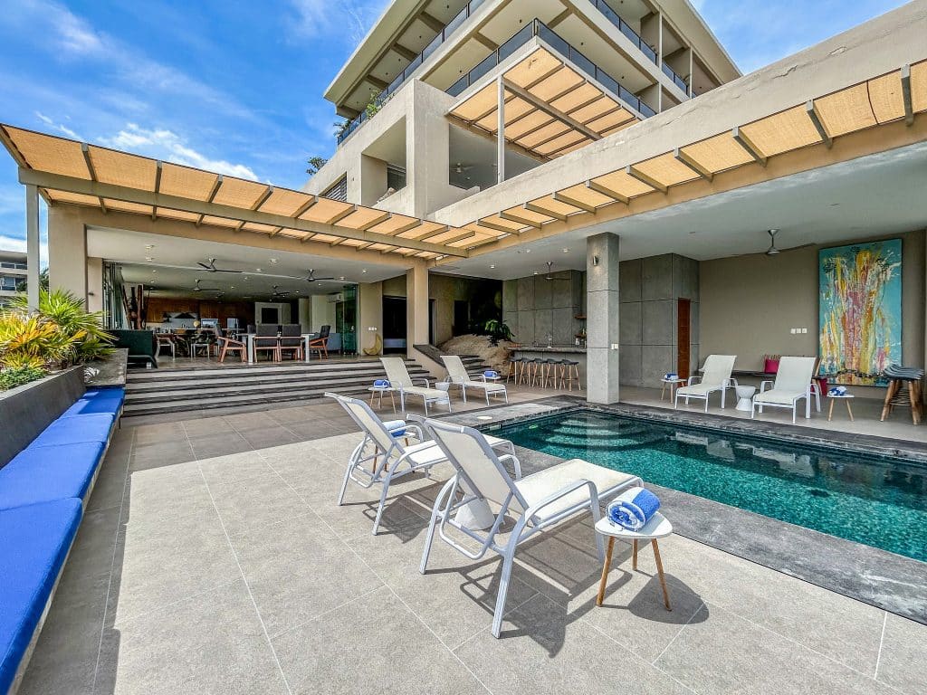 Outdoor view of Villa Montecarlo in Huatulco showing a private pool area with lounge chairs, a covered patio with dining and living spaces, and modern architecture against a blue sky.
