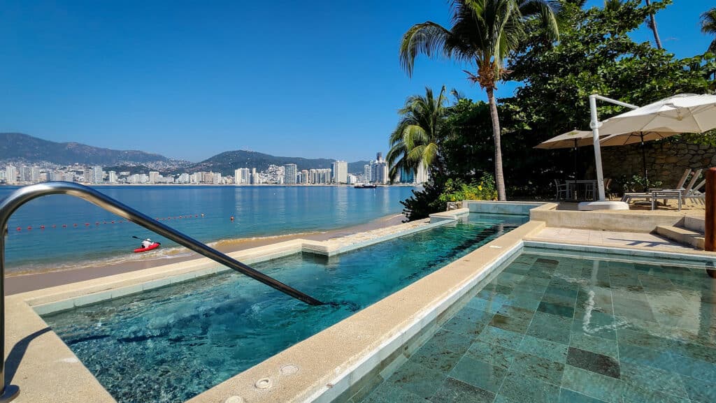 Beachfront pool and lounge area at Villa Alejandra Garden 1000 in Acapulco, with city skyline and mountains in the background