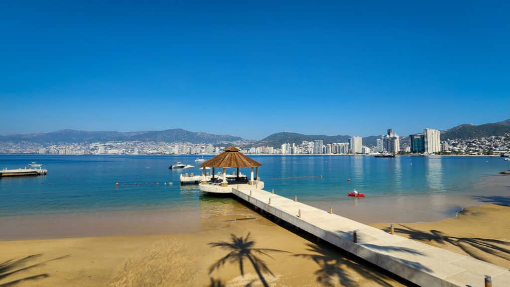 Beachfront view from Villa Alejandra Garden 1000 in Acapulco, featuring a pier with a gazebo, sandy beach, and city skyline