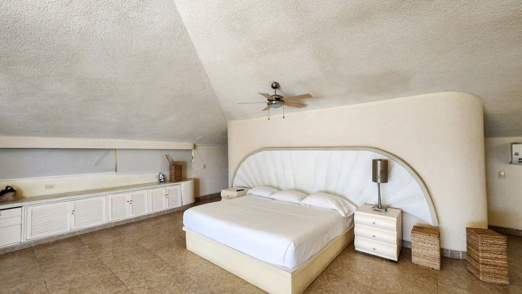 Interior of Lomas Del Marques House in Acapulco showing a white bed, nightstand, wicker stools, and a ceiling fan