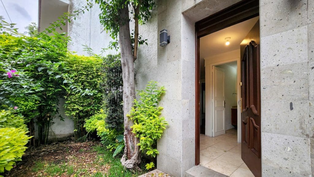 Entrance of a Milton 64 home in Miguel Hidalgo, Mexico City, with a wooden door, green plants, and a small tree beside the doorway.