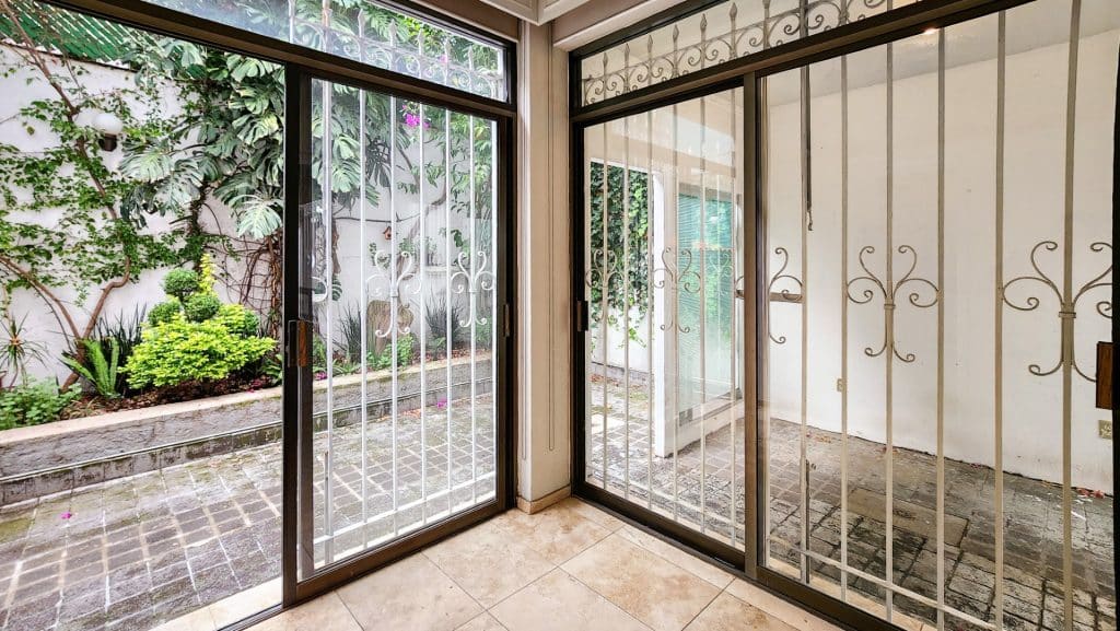 View of a small outdoor garden with green plants and flowering bushes, seen through metal security doors in a room at Milton 64 in Mexico City.