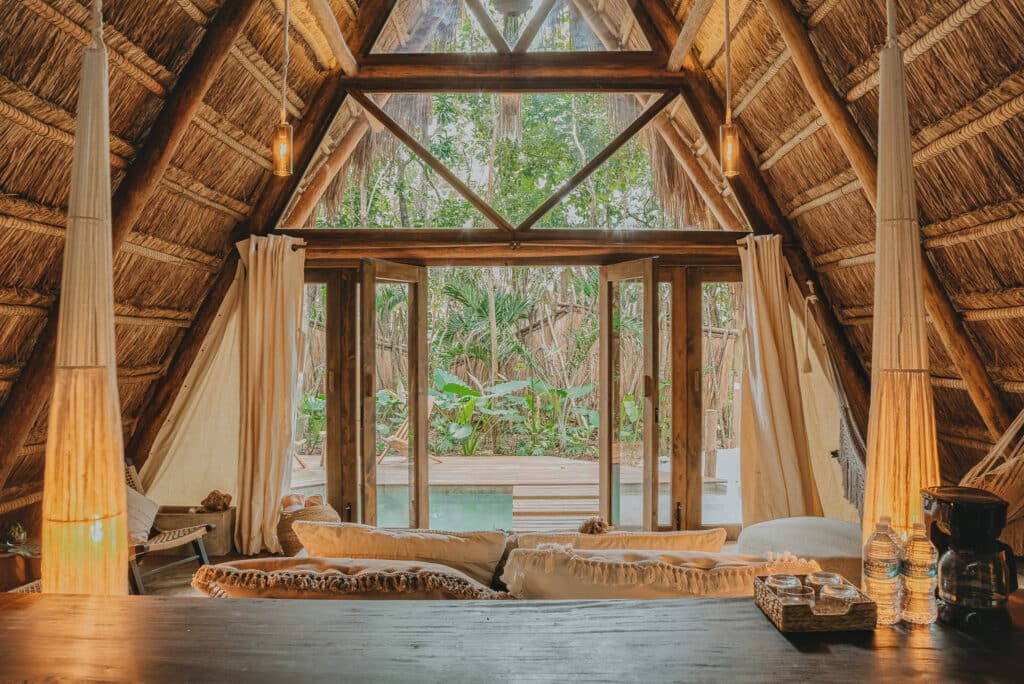 Interior view of a thatched-roof cabin at 13 in Lake Front Bacalar, showing open doors to a lush outdoor area with a small