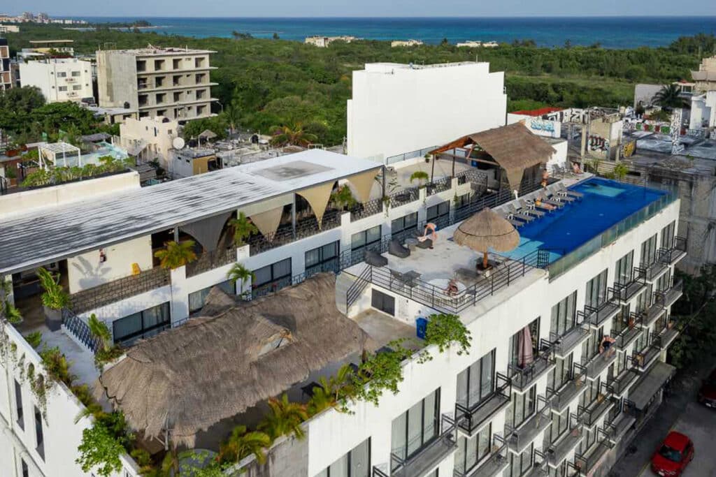 Aerial view of the 82 Room Playa del Carmen Hotel in Playa del Carmen, showing its rooftop pool, terraces with sun loungers, shaded seating areas, and surrounding greenery and buildings.