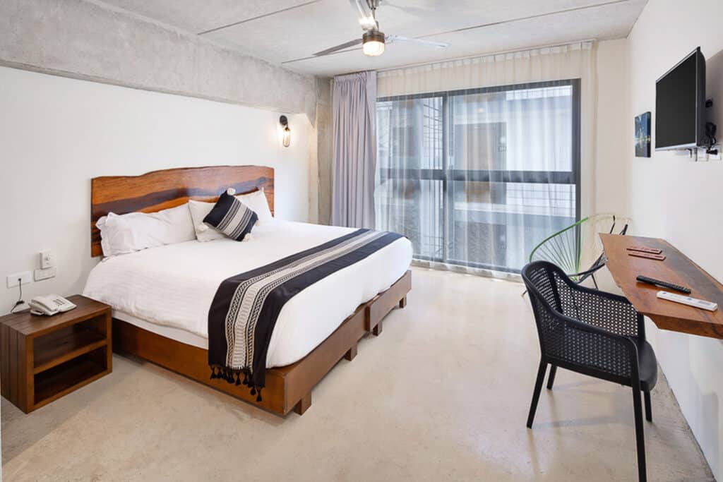 A modern hotel room at 82 Room Playa del Carmen in Playa del Carmen, featuring a king-sized bed with wooden accents, a wall-mounted TV, and a large window with sheer curtains.