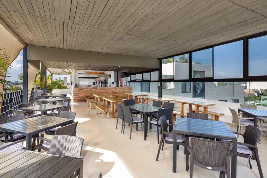 A spacious indoor dining area at 82 Room Playa del Carmen Hotel in Playa del Carmen, featuring wooden and wicker furniture, large windows, and a view of nearby buildings.