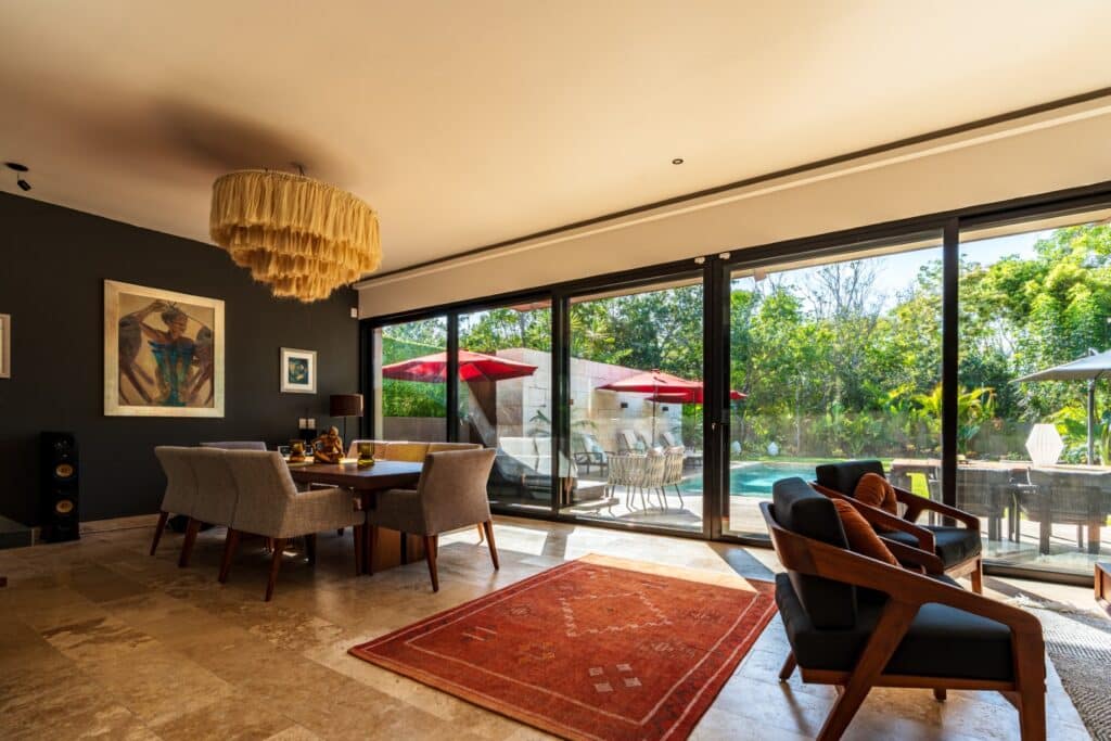 Interior of a villa at Brisas 45 in Playa del Carmen, featuring modern furniture, a dark accent wall with artwork, and large sliding glass doors opening to a pool area with red umbrellas.
