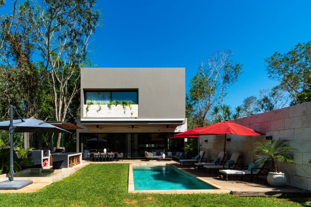 A modern two-story villa in Brisas 45 – Villa in Xpu Ha, Playa del Carmen, features a lawn, a swimming pool, outdoor lounge areas with umbrellas, and lush greenery under a blue sky.