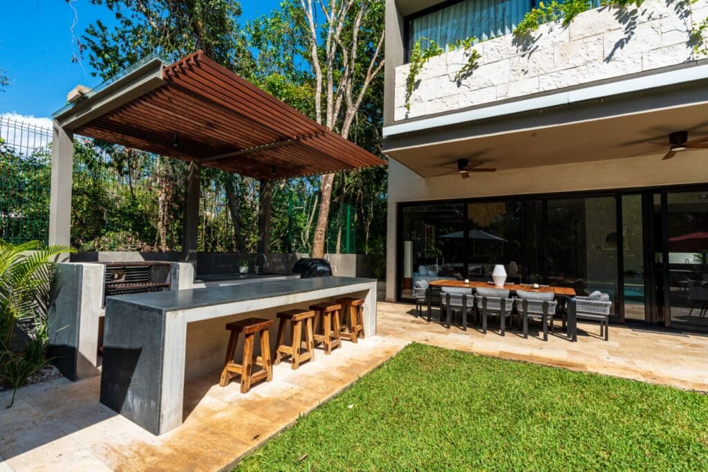 A modern villa with an outdoor kitchen and dining area, situated in the Brisas 45 development in Playa del Carmen's Xpu Ha area, surrounded by lush greenery.