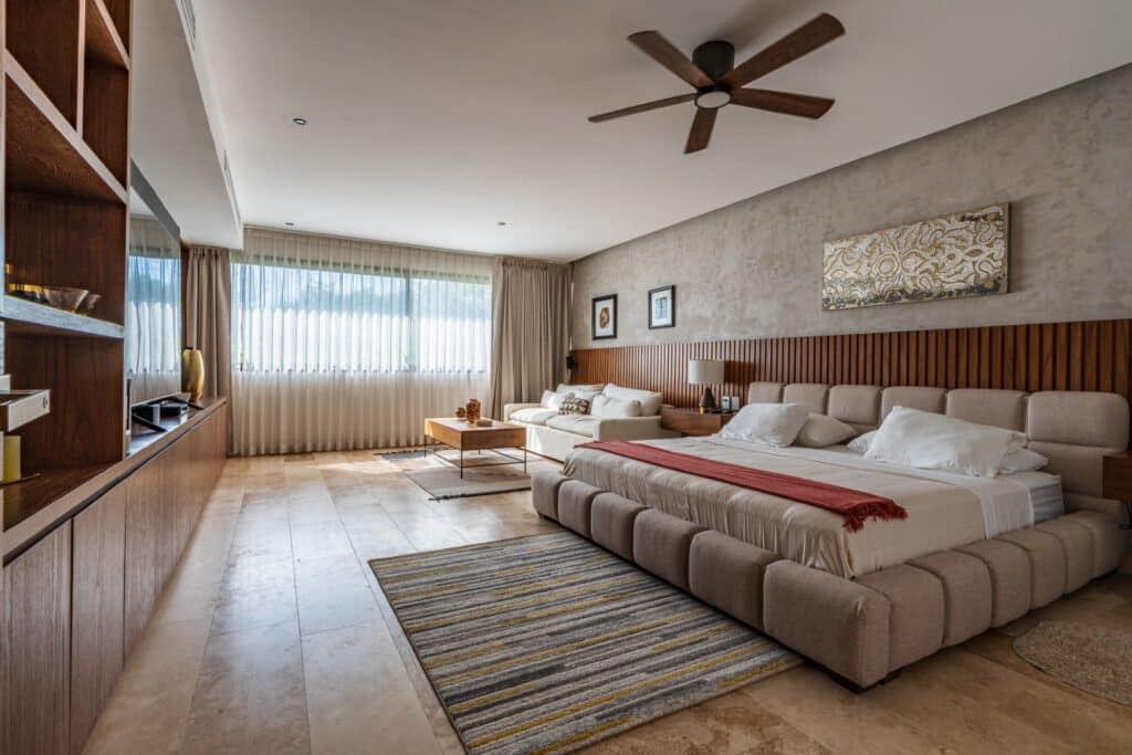 Living area of a villa in Brisas 45 in Xpu Ha, Playa del Carmen, featuring a beige tufted bed, white sofa, wooden coffee table, large window with curtains, and ceiling fan.