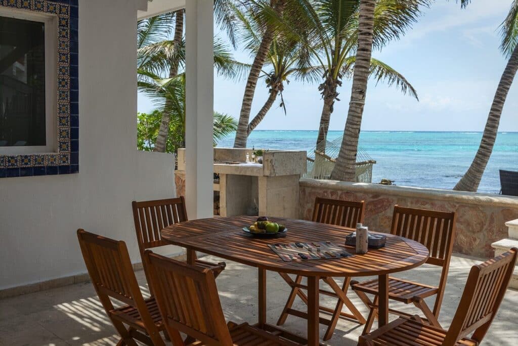 A view from Casa Alea in Tankah Bay #29 showing a wooden dining table with six chairs on a shaded patio overlooking palm trees and the turquoise Caribbean Sea in Tulum.