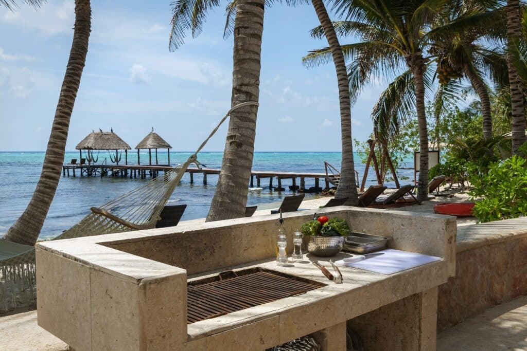 A beachfront scene at Tankah Bay #29 in Tulum, featuring a stone outdoor kitchen with a view of palm trees, lounge chairs, and a small pier over clear turquoise water.