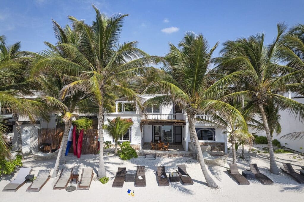 A beachfront house at Tankah Bay #29 in Tulum features a white exterior, surrounded by tall palm trees, with lounge chairs on the sandy beach and a shaded patio area.