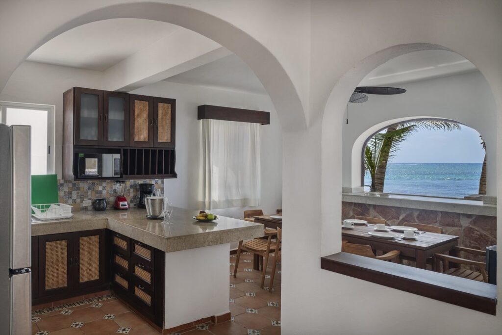 Interior view of Casa Alea at Tankah Bay #29 in Tulum, showcasing a kitchen with wooden cabinets and a dining area with ocean views through arched openings.