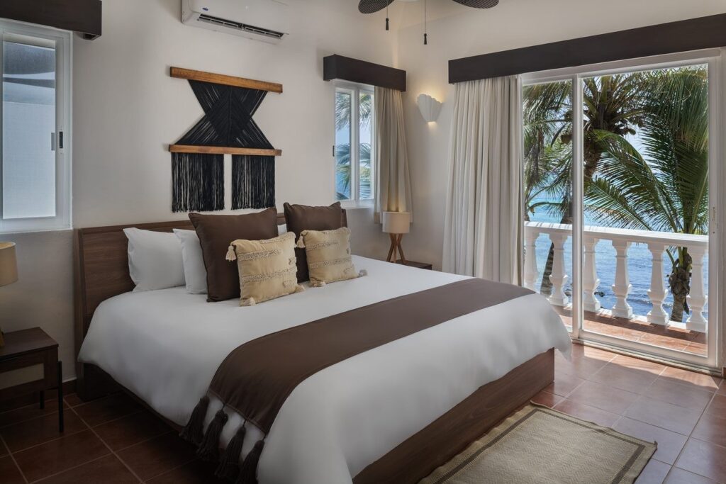 A bedroom with a wooden bed frame, beige and brown cushions, and a view of the ocean and palm trees through sliding glass doors at Tankah Bay #29 in Tulum.