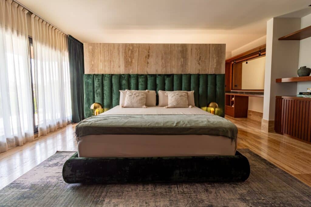 A stylish bedroom in Flamingos Villa located in Cancun's Marina Front area, featuring a green velvet headboard, wooden accents, and large windows with sheer curtains.