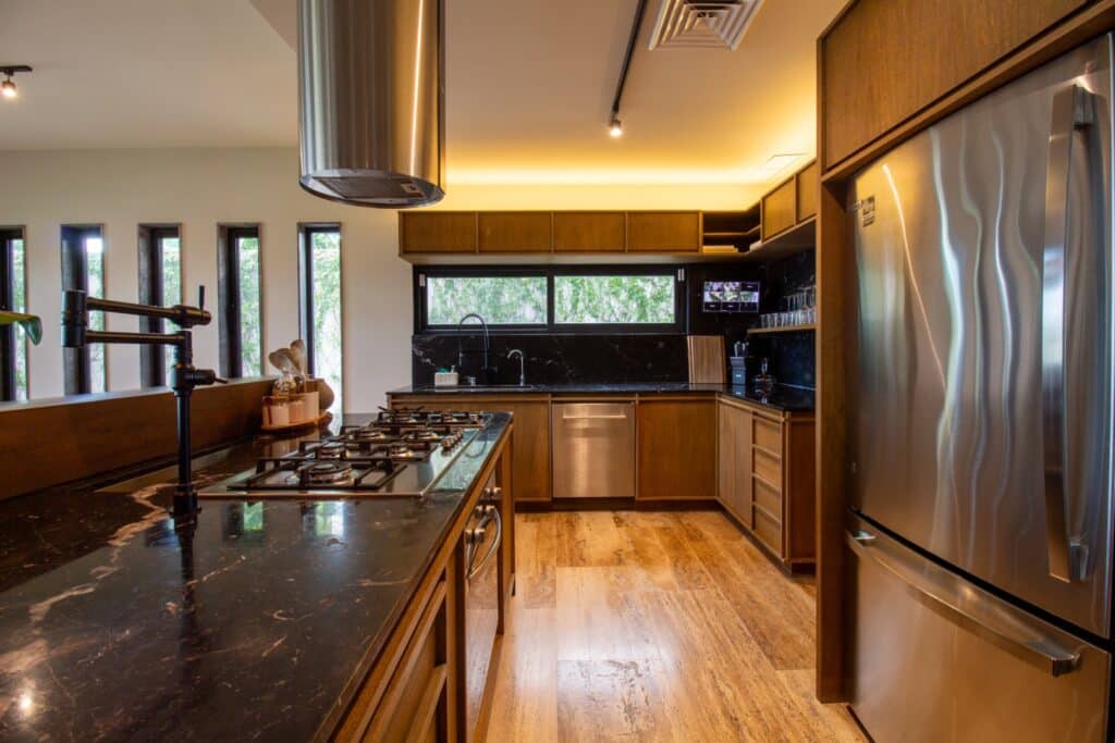 Modern kitchen in Villa Flamingos Cancun located in the Marina Front area, featuring wooden cabinets, a black marble countertop, stainless steel appliances, and ample natural light.