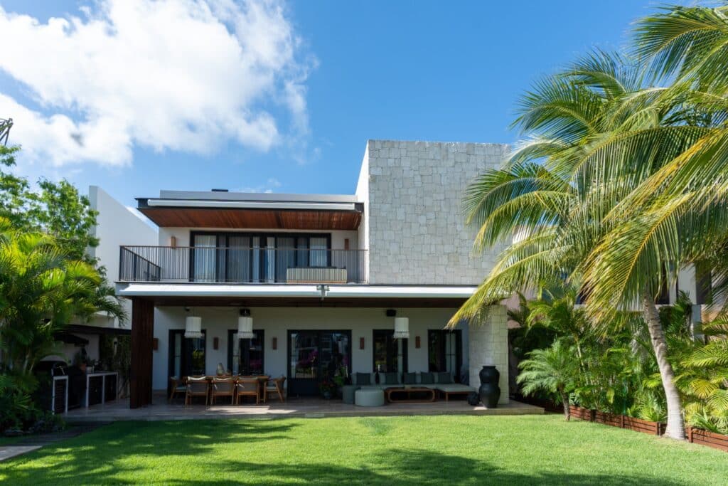 A modern two-story villa in Flamingos Villa at Marina Front in Cancun features a spacious outdoor patio with lush green lawn and tropical palm trees under a blue sky.