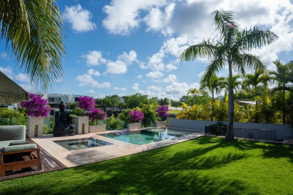 A view of Villa Flamingos in Cancun's Marina Front area, featuring a small pool surrounded by lush greenery, palm trees, vibrant purple flowers, and a blue sky with scattered clouds.