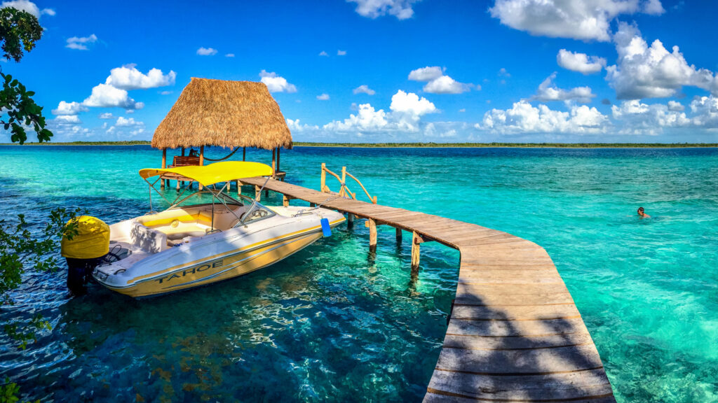 A luxury lakefront estate villa in Bacalar features a wooden dock extending over clear turquoise waters, a white boat with yellow accents, and a thatched-roof hut under a bright blue sky.