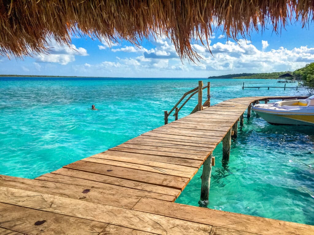 A wooden dock extends over clear turquoise waters at Bacalar, with a thatched roof overhead and a person swimming in the lake, surrounded by a lush green shoreline.