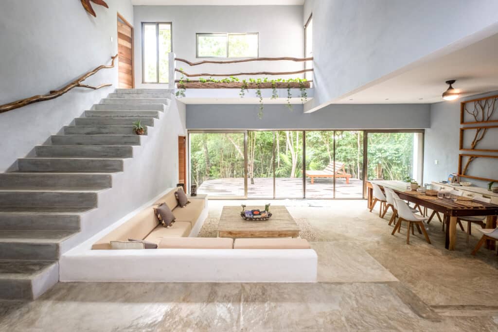 A spacious, modern living area in Bacalar development, featuring large sliding glass doors opening to lush greenery, a cozy built-in corner sofa, and a minimalist wooden dining table.
