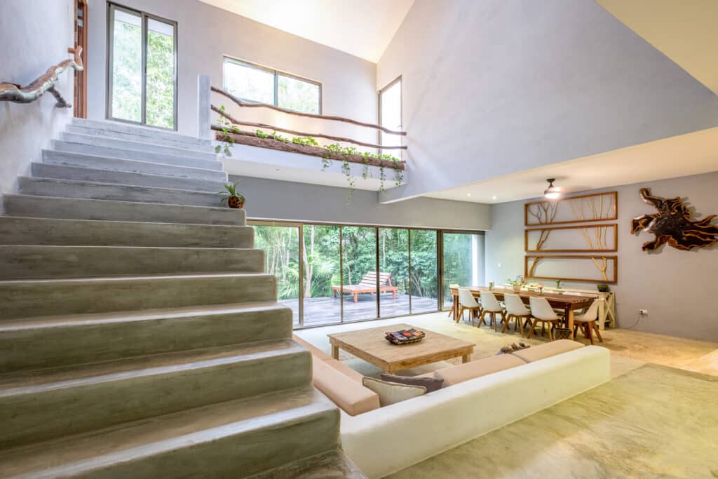 Interior view of the Bacalar luxury lakefront estate villa, featuring a spacious living area with large windows, a concrete staircase, and modern decor overlooking greenery.