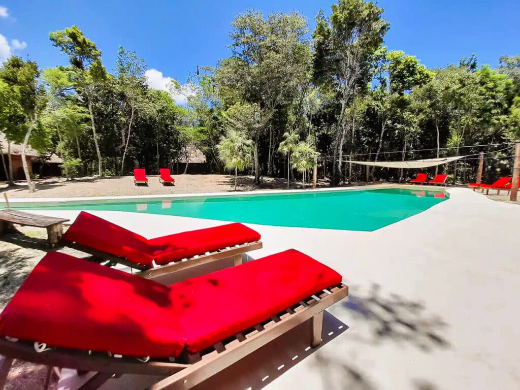 A view of the pool area at 10 Cabin Jungle Retreat in Puerto Aventuras, with red lounge chairs, surrounded by trees and a clear blue sky overhead.