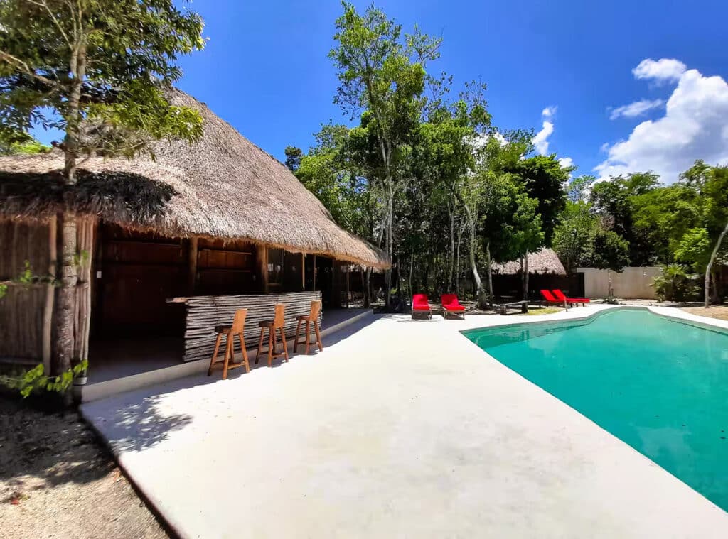 A jungle retreat hotel in Xpu Ha features a thatched-roof cabana, a swimming pool, and red lounge chairs surrounded by lush trees under a bright blue sky in Puerto Aventuras.