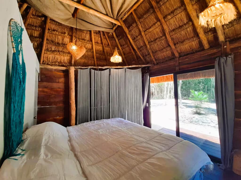 A cozy bedroom in the 10 Cabin Jungle Retreat with wooden walls and a thatched roof, overlooking a lush outdoor area through large sliding glass doors., Puerto Aventuras.