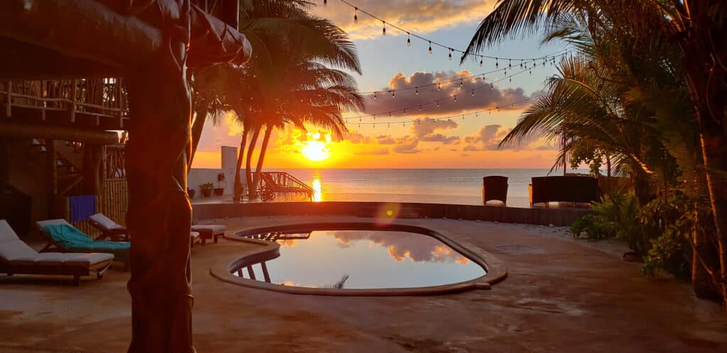 A beachfront villa in Villa Sirena, Puerto Morelos, with a pool overlooking the ocean during a sunset, surrounded by palm trees and outdoor lounge chairs.