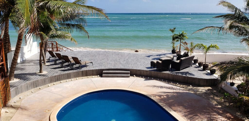 A beachfront terrace at Villa Sirena in Puerto Morelos features lounge chairs, cozy seating, and lush palm trees overlooking the ocean and a small pool in the foreground.