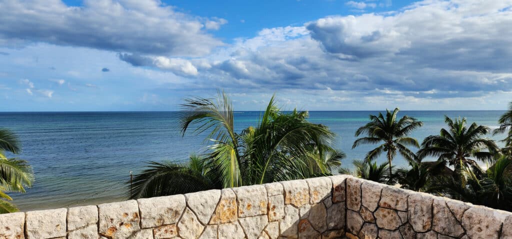 View from Villa Sirena in Puerto Morelos overlooking the beachfront with palm trees and the ocean under a partly cloudy sky.