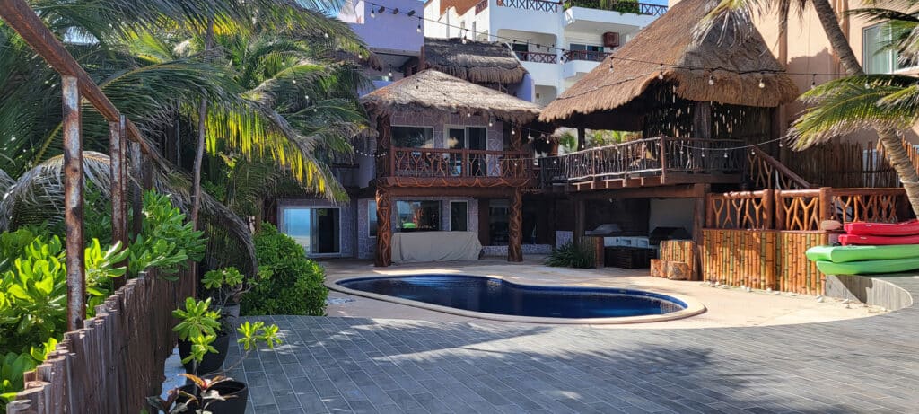 A beachfront villa at Villa Sirena in Puerto Morelos features a small pool, tropical plants, and a thatched-roof deck surrounded by lush greenery.