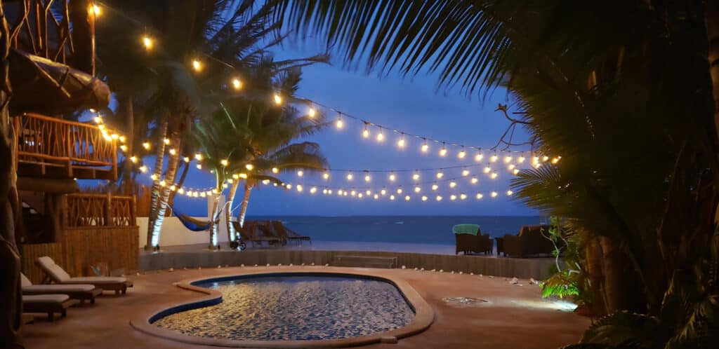 A view of Villa Sirena in Puerto Morelos featuring a beachfront pool area with lounge chairs, string lights overhead, and the ocean in the background at dusk.