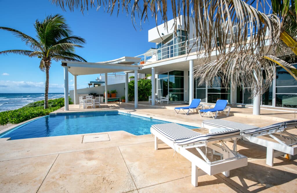 A beachfront house at Dos Bahias in Tulum features a private pool, outdoor lounge area, and floor-to-ceiling windows overlooking the ocean under a clear blue sky.