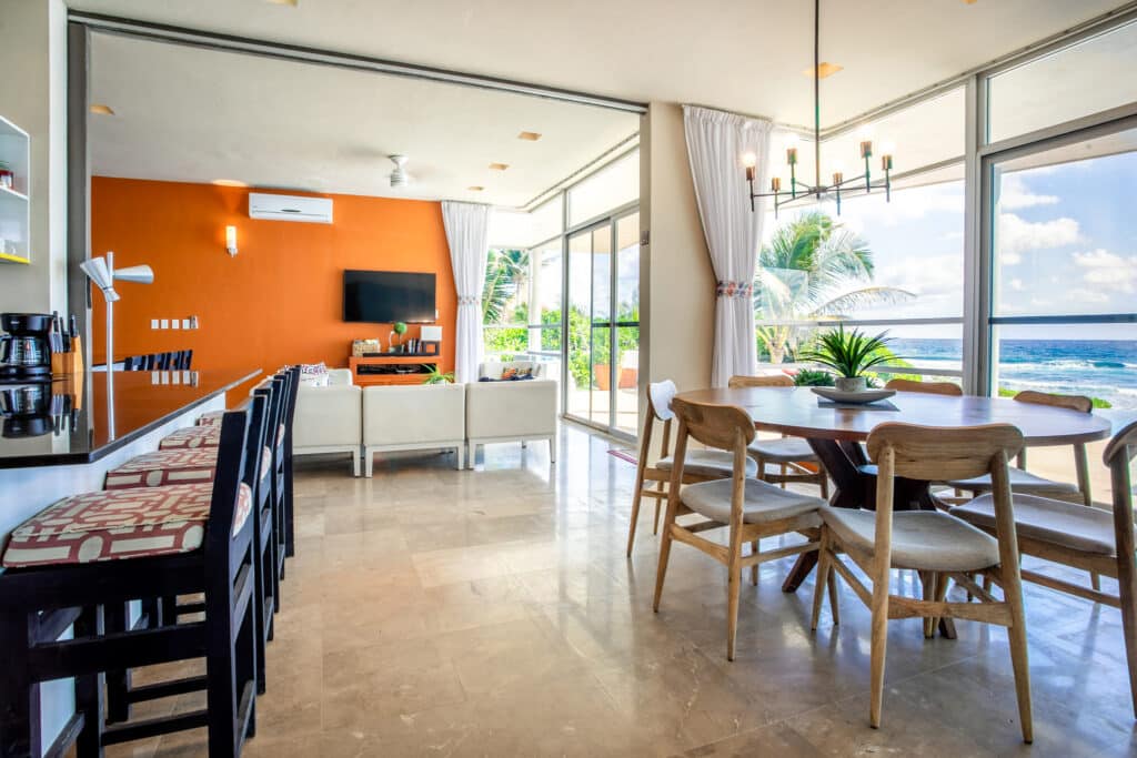 Living area of a beachfront house at Dos Bahias in Tulum, featuring large windows with ocean views, a wooden dining table, and a modern open-concept design.