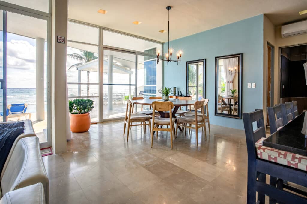 Interior view of a beachfront house at Dos Bahias in Tulum, featuring a dining area with a round wooden table, natural light, and ocean views through large glass doors.