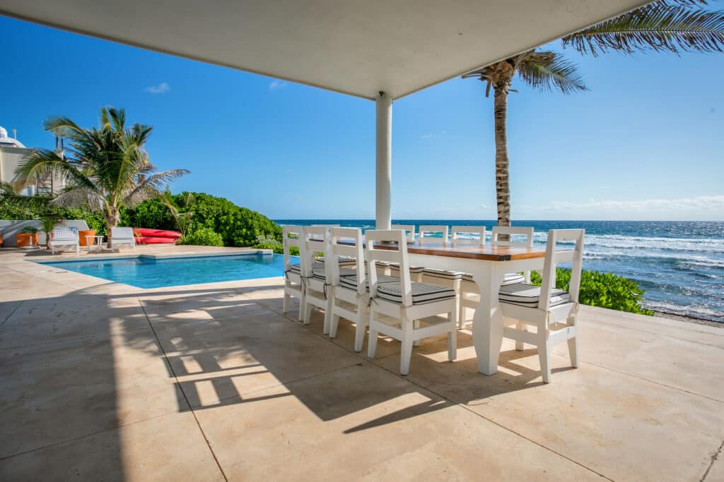 A beachfront house in Dos Bahias, Tulum, with a covered patio, white dining table and chairs, a swimming pool, palm trees, and ocean views.