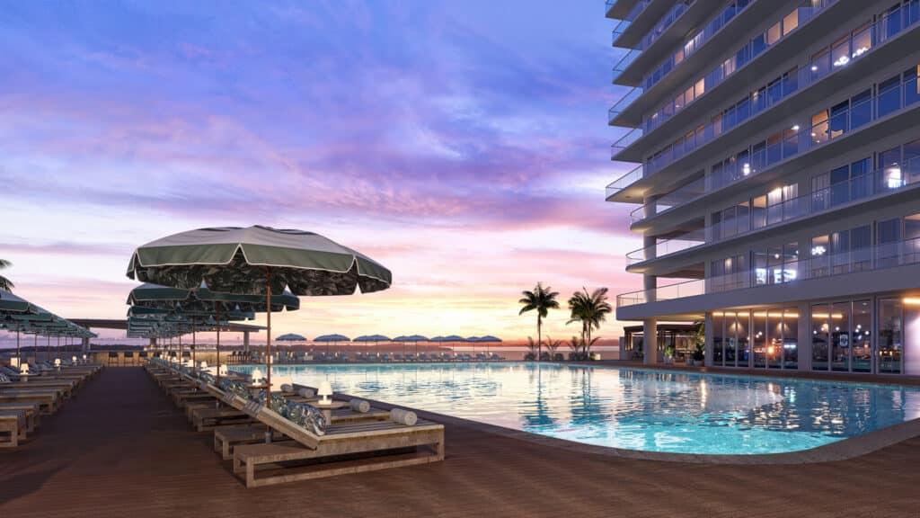 A sunset view at Bay View Grand 1419-B in Cancun features a pool surrounded by lounge chairs and umbrellas, with a modern building and palm trees in the background.