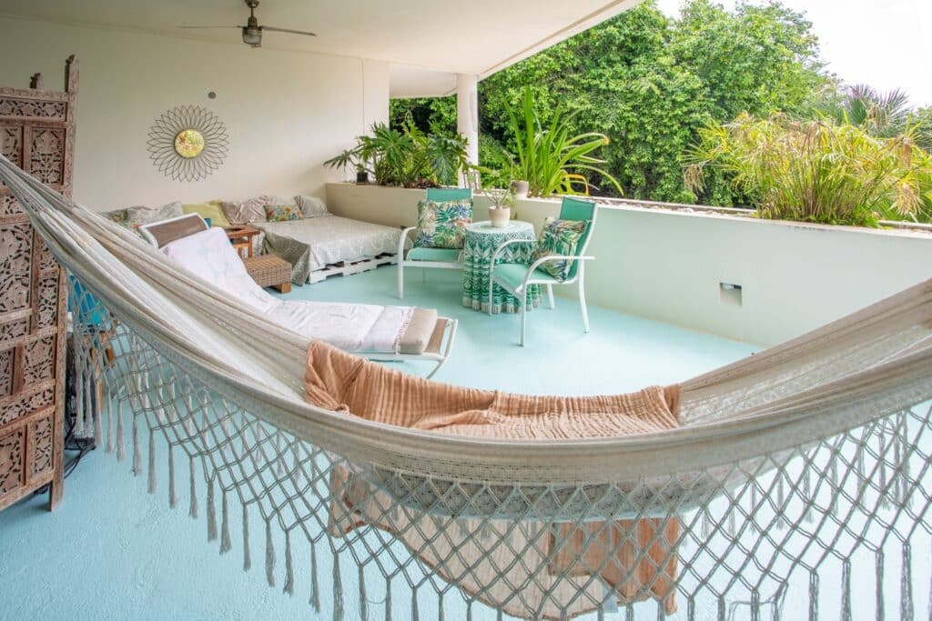 A spacious balcony at Bosque Los Aluxes Condo in Playa del Carmen features a hammock, outdoor seating with a table, lush greenery, and an inviting view of tropical plants.